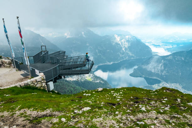 Woman at the 5 Fingers observation platform on top of the Krippenstein mountain, enjoying the stunning view of the Salzkammergut region, OÖ, Austria Krippenstein, Austria - July 16th 2019: From the 5 Fingers, a steel construction on top of the Krippenstein Mountain at the Dachstein glacier massif, tourists and hikers can enjoy an unique and amazing view of the Salzkammergut region with it's beautiful lakes and mountains. Clouds at the same altitude as this observation point give the picture a dramatic note. Down at the valley, the Lake Hallstatt is visible, with the famous mountain village Hallstatt at it's shore. 
A woman stands alone at the platform and enjoys the thrilling view. dachstein mountains stock pictures, royalty-free photos & images