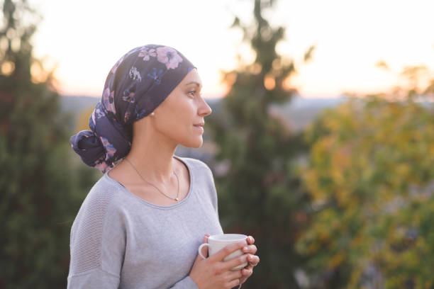 woman battling cancer stands outside and contemplates her life A beautiful young woman wearing a head wrap looks off in the distance and smiles contemplatively while drinking a cup of tea. She is standing outdoors and there are mountains and trees in the background. patience stock pictures, royalty-free photos & images