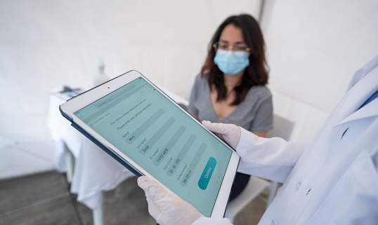 Young woman being registered for her COVID-19 vaccine at an immunization stand using a tablet computer. **DESIGN ON SCREEN WAS MADE FROM SCRATCH BY US**