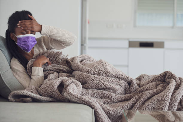 Woman checking fever. Young ill woman holding hand on forehead, checking temperature, resting, lying on the couch with a cozy blanket. Using purple face mask to prevent other people from getting infected. cold and flu stock pictures, royalty-free photos & images