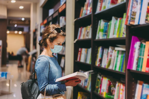Woman choosing a new book in the bookstore during COVID-19 pandemic Woman with protective face mask reading a book in the bookstore bookstore stock pictures, royalty-free photos & images