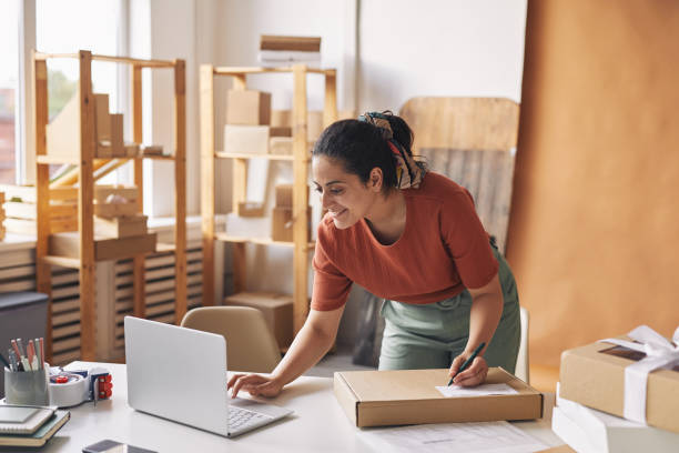 Woman completing orders by laptop Young smiling woman typing on laptop and accepting the orders then packing parcels for shipment small business stock pictures, royalty-free photos & images