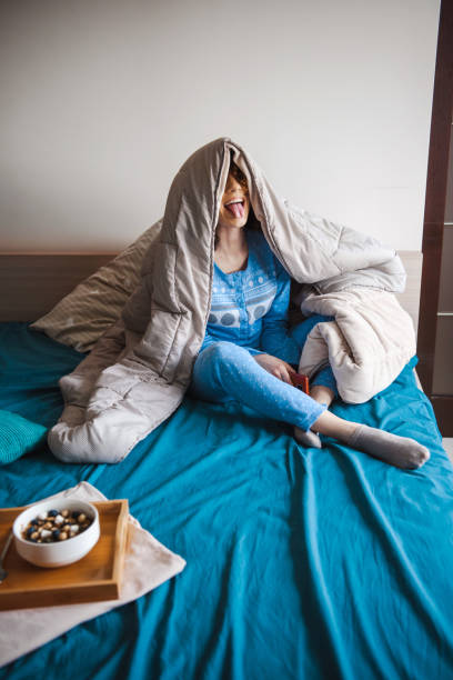 Woman covering her head with a blanket sitting in bed while grimacing. Morning time. Healthy eating. stock photo