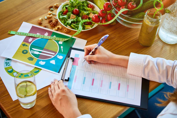 Woman dietitian in medical uniform with tape measure working on a diet plan sitting with different healthy food ingredients in the green office on background. Weight loss and right nutrition concept stock photo