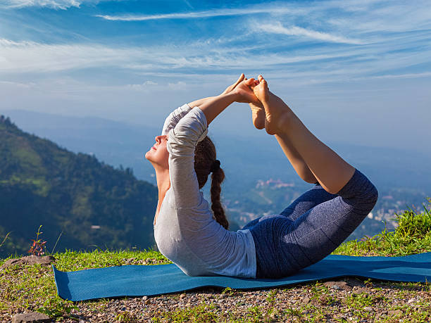 Woman doing Ashtanga Vinyasa Yoga asana Dhanurasana - bow pose Yoga outdoors - young sporty fit woman doing Ashtanga Vinyasa Yoga asana Dhanurasana - bow pose  - in Himalayas mountains in the morning  Himachal Pradesh, India flexibility workout stock pictures, royalty-free photos & images