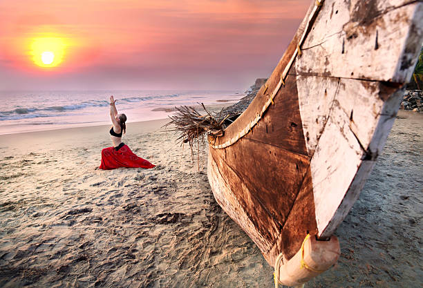 Woman doing warrior yoga pose by a boat on a beach at sunset stock photo