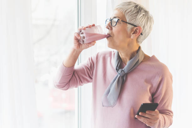 Woman drinking healthy smoothie Beautiful senior woman with modern short gray hair and glasses drinking healthy fresh fruit smoothie by the window in her bright home and holding mobile phone in her hand, Healthy modern and natural lifestyle concept drinking smoothie stock pictures, royalty-free photos & images