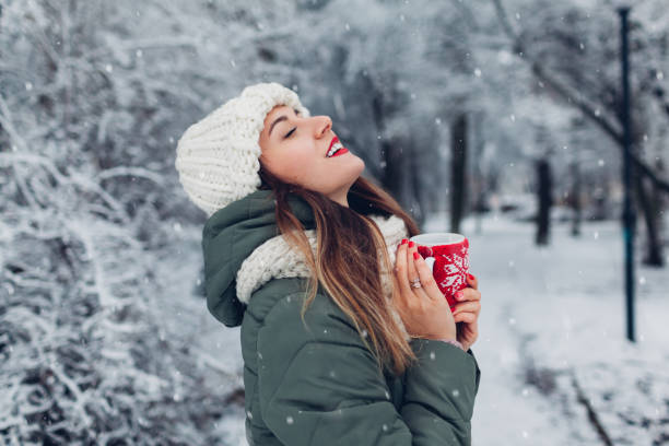 Woman drinking hot tea holding vacuum flask in snowy winter park. Cup dressed in red knitted Christmas case stock photo
