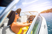 Young and pretty woman in yellow skirt and swimsuit with hat and sunglasses driving luxury yacht in the sea. 