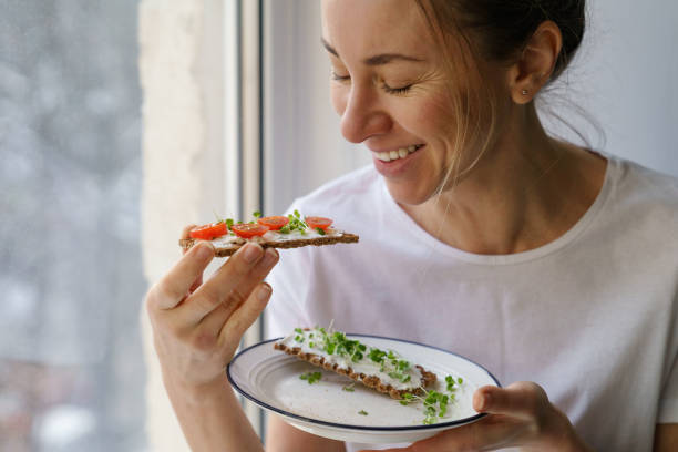 Woman eating rye crisp bread with creamy vegetarian cheese tofu, tomato, micro greens. Healthy food Smiling woman eating rye crisp bread with creamy vegetarian cheese tofu, cherry tomato and rucola micro greens, sitting at home and looking at window. Healthy food, gluten free, diet concept. crucifers stock pictures, royalty-free photos & images