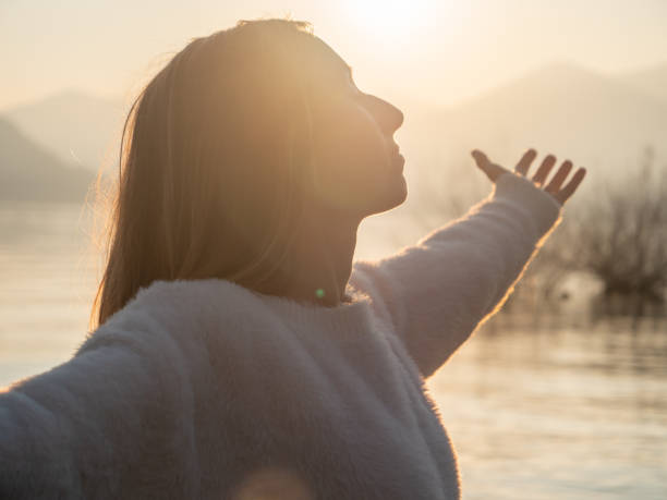 Woman embraces nature, she stands arms out by the lake at sunset stock photo