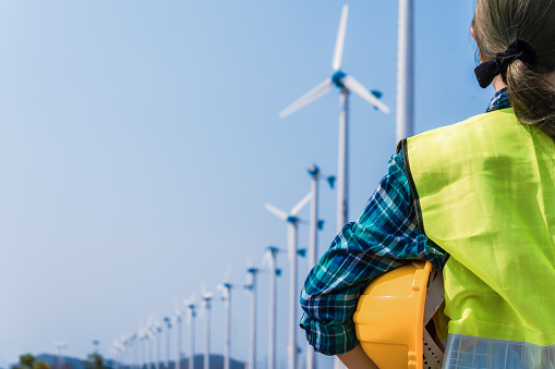 Woman engineer in uniform and holding yellow safety helmet with standing and checking wind turbine power in construction site renewable energy. Clean energy and environment concept.