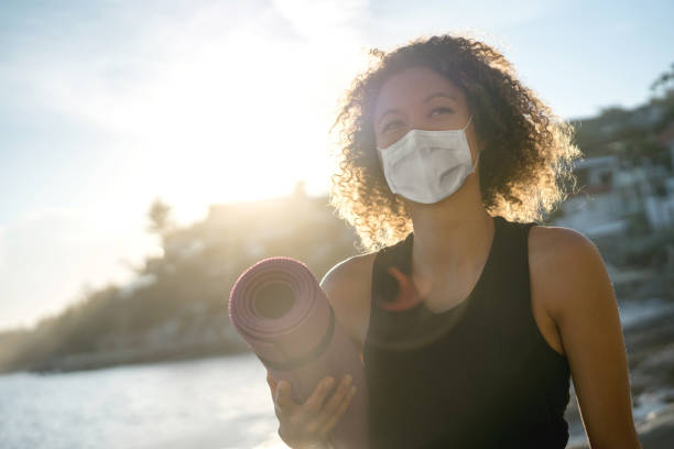 mulher se exercitando na praia usando uma máscara facial e segurando um tapete de ioga - cuidado com o corpo - fotografias e filmes do acervo