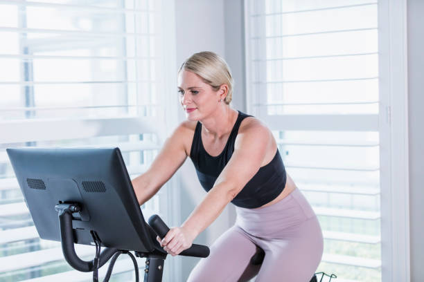 Woman exercising on bike at home A mid adult woman in her 30s working out at home, on an exercise bike. peloton stock pictures, royalty-free photos & images