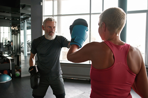 Back view middle age woman with short blond hair wearing pink top and blue boxing gloves hits trainer into punch mits. Mature professional trainer helps woman to learn boxing techniques.