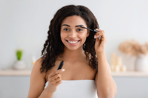Woman getting ready for work, doing morning makeup routine in bathroom at home. Glad millennial black woman in white towel applies mascara with brush on eyelashes in bright interior, empty space