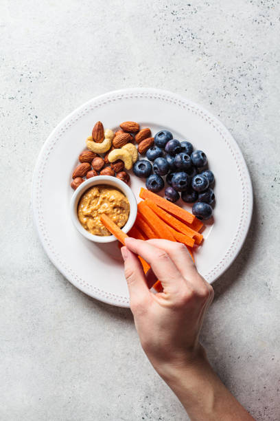 Woman hand eating healthy snacks plate - carrot, nut, berry and peanut butter, top view. stock photo