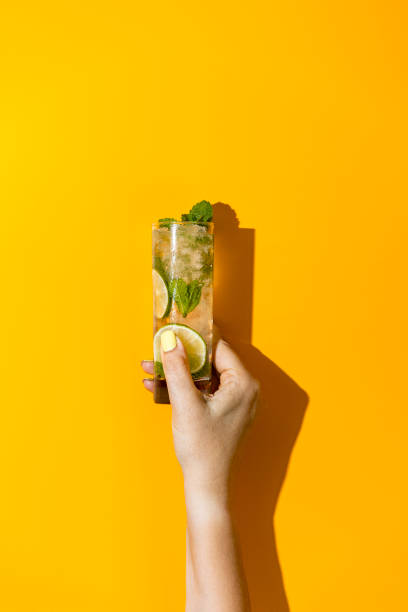 Woman hand hold glass with water drops of Mojito cocktail drink with lime Ice and mint on bright yellow background. Summer art food concept on yellow background. stock photo