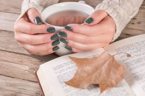 Woman hands in sweater with olive color manicure holds cup of tea and open book stock photo