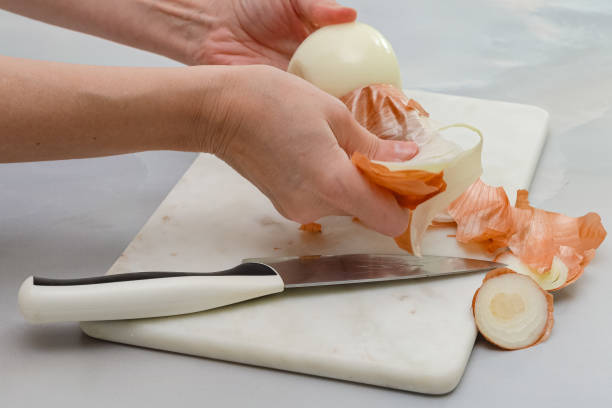A woman hands peeling white onion with knife, close up view, marble kitchen table background stock photo