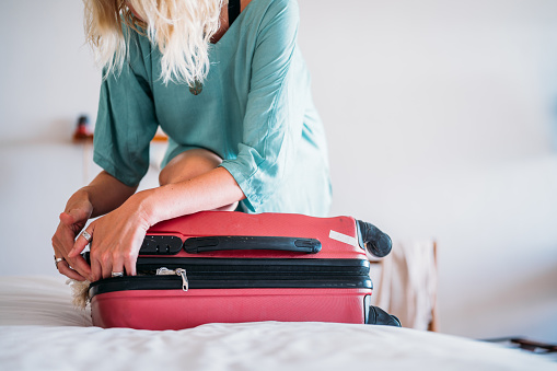 Close-up of a woman in a turquoise dress, trying to close a full suitcase with her knee.