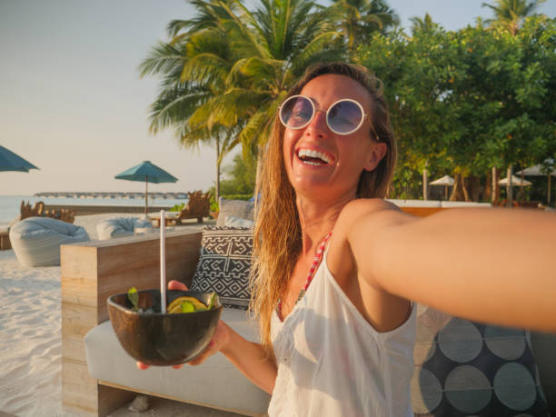 Woman having cocktail at sunset on tropical beach and taking selfie stock photo