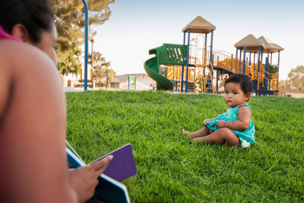 A woman holding a childrens book tries to engage the attention of a baby girl sitting in the lawn of a kids playground. A woman holding a childrens book tries to engage the attention of a baby girl sitting in the lawn of a kids playground. hot latino girl stock pictures, royalty-free photos & images