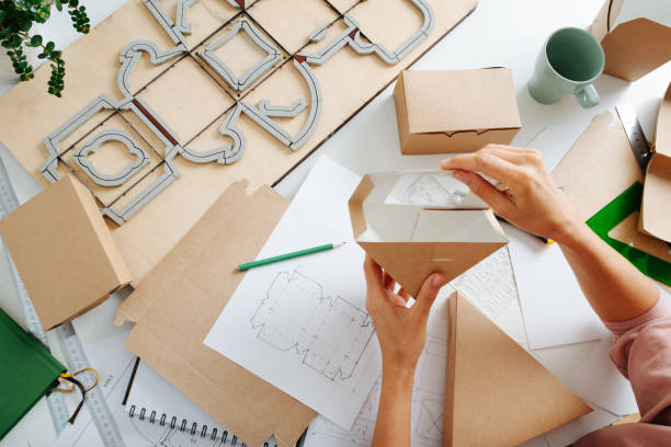 Woman holding box with a hole above her workplace Female box maker working behind a table in a private workshop, folding, shaping a box, tools scattered across. Rulers, pencils and special board with a cutting form. packaging stock pictures, royalty-free photos & images