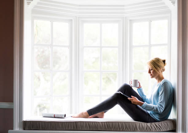 Woman holding coffee and digital tablet by window Young woman having coffee and looking at digital tablet. Full length of female sitting on alcove window seat. She is at home on sunny day. alcove window seat stock pictures, royalty-free photos & images