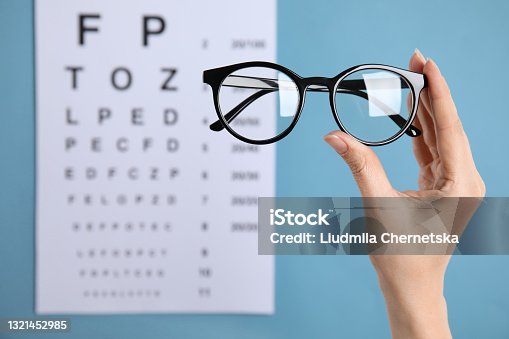 istock Woman holding glasses against eye chart on blue background, closeup. Ophthalmologist prescription 1321452985