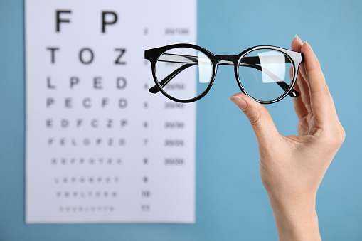 Woman holding glasses against eye chart on blue background, closeup. Ophthalmologist prescription