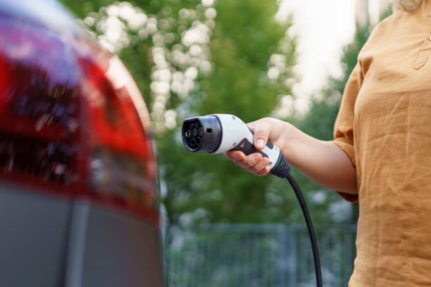 Woman holding power supply cable at electric vehicle charging station, closeup stock photo