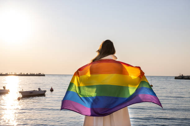 Woman holding pride flag facing the sun looking at the sea. stock photo