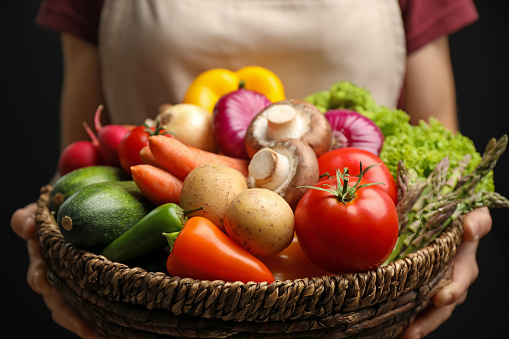 Woman holding wicker basket full of fresh vegetables on black background, closeup