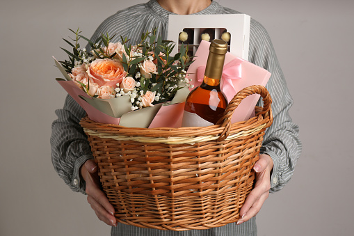 Woman holding wicker basket with different gifts on grey background, closeup