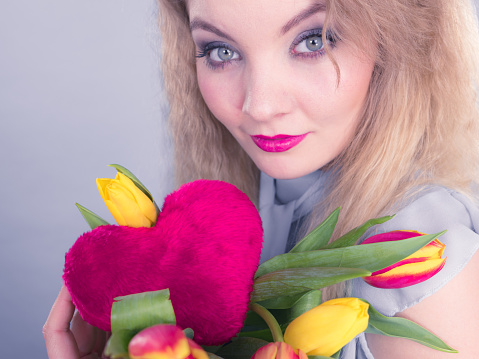 International womens or valentines day. Attractive grateful woman blonde hair holding tulips bunch and red heart sign. On grey