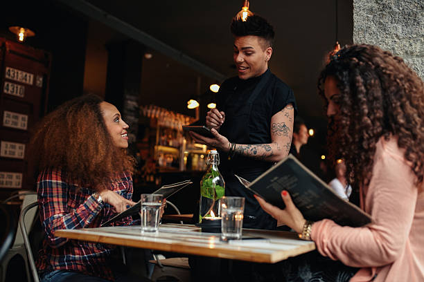 Woman in a cafe ordering to waiter Young woman with a friend ordering to waiter holding digital tablet. Two women sitting at cafe holding menu card giving an order to male waiter. waiter taking order stock pictures, royalty-free photos & images