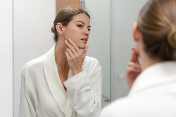 Woman in bathrobe looking in mirror at bathroom Young and gorgeous woman in white bathrobe standing in bright light bathroom with mirror. She looking at her reflection with calm face, holding hand on cheek, checking skin complexion stock pictures, royalty-free photos & images