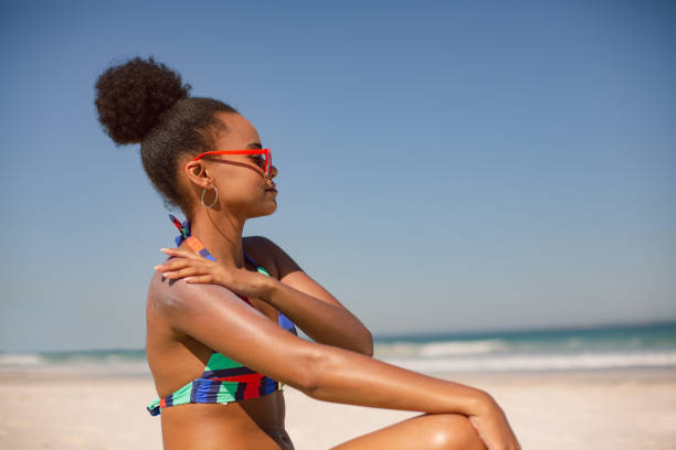 frau im bikini auftragen sonnencreme lotion auf schulter am strand in der sonne - sonnencreme stock-fotos und bilder