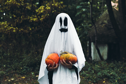 Woman dressed like a spooky smiling ghost holding two beautiful orange pumpkins and staying in the scary forest near abandoned house celebrating Halloween