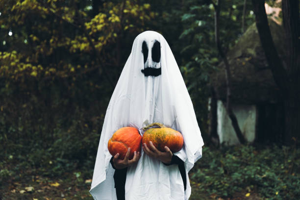 Woman in ghost costume with pumpkins wish you a Happy Halloween! Woman dressed like a spooky smiling ghost holding two beautiful orange pumpkins and staying in the scary forest near abandoned house celebrating Halloween stage costume stock pictures, royalty-free photos & images