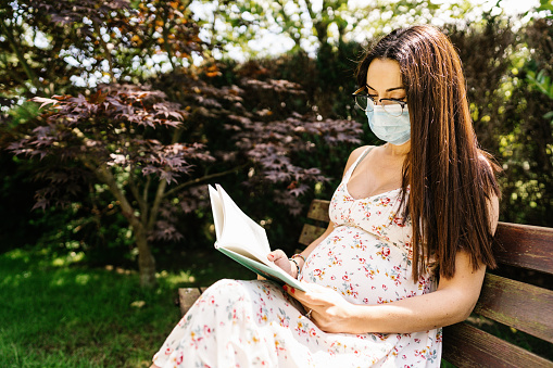 Female in floral dress and medical mask sitting on bench and reading book during epidemic on sunny summer day in park