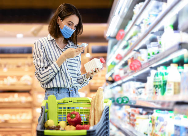 Woman in mask scanning code on products using smartphone Young woman in disposable face mask taking dairy products from shelf in the supermarket, holding bottle and smartphone, scanning bar code on product through mobile phone, walking with trolley cart aisle photos stock pictures, royalty-free photos & images