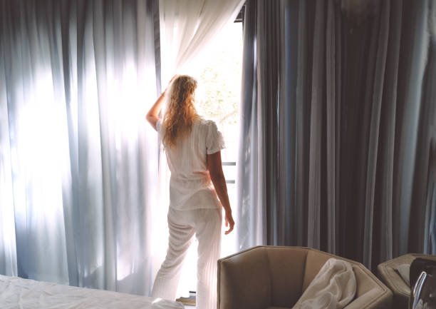 Woman in white pajamas opening window curtains in the morning stock photo
