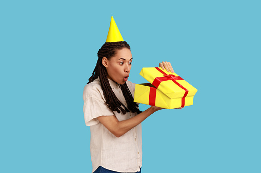 Portrait of surprised woman in yellow party cone looking into gift box, opening present and peeking inside with shocked expression, wearing white shirt. Indoor studio shot isolated on blue background.