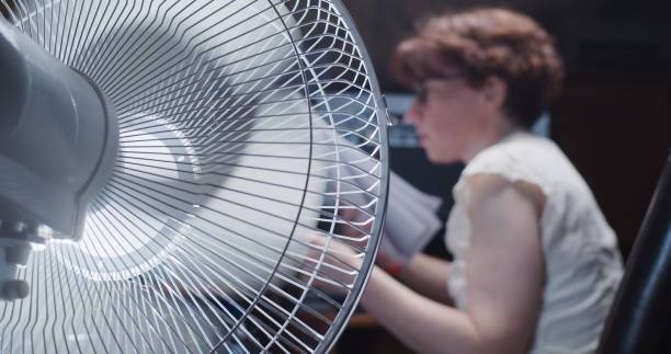 Woman is trying to work at the desktop computer at home, suffering from hot weather and using electric fan stock photo