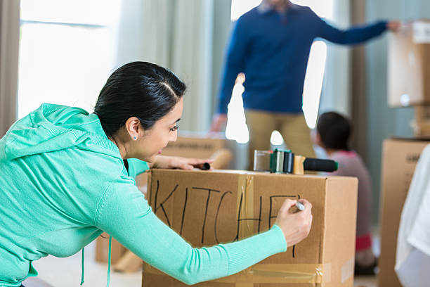 Woman labels box with kitchen supplies Mid adult Hispanic woman writes 'kitchen' with a marker. She has just packed up some of the kitchen supplies as she and her family prepare to move. Her husband and daughter are working in the background. labeling stock pictures, royalty-free photos & images