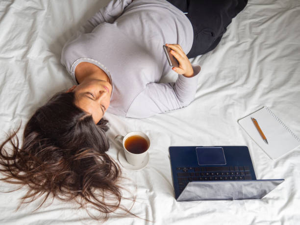Woman laying in bed looking at cellphone procrastinating work. stock photo