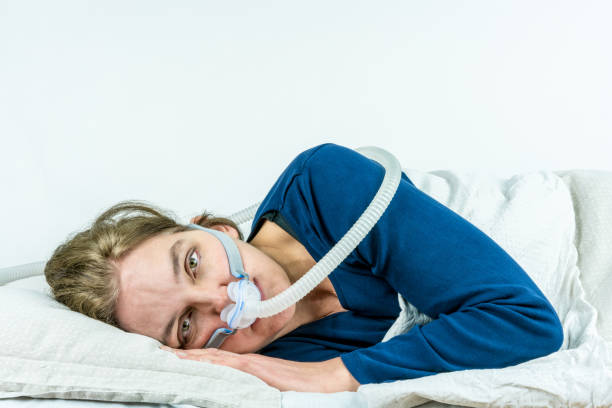 Woman laying on her side with eyes open looking in to camera. CPAP, sleep apnea treatment. Studio portrait white background. stock photo