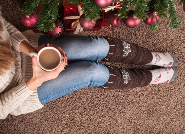 Woman legs with warmers on floor on carpet with cup of coffee in hand and next to Christmas tree stock photo